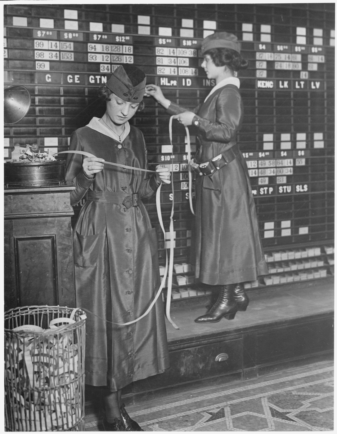 Two women read ticker tape and post prices on a quotation board, 1918